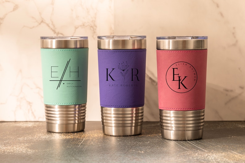 May include: Three stainless steel tumblers with personalized leather wraps. The tumblers are in the colors teal, purple, and pink. The teal tumbler has the initials "E/H" and the name "EMIL HUTCHINSON". The purple tumbler has the initials "KVR" and the name "KATE ROBERTS". The pink tumbler has the initials "EK" and the name "ELIZA KATHMAN".