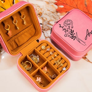Puede incluir: Caja de joyer&iacute;a de terciopelo rosa con un dise&ntilde;o floral y el nombre "Anna" grabado en la tapa. La caja est&aacute; abierta y llena de varias piezas de joyer&iacute;a, incluyendo anillos, aretes y un collar.