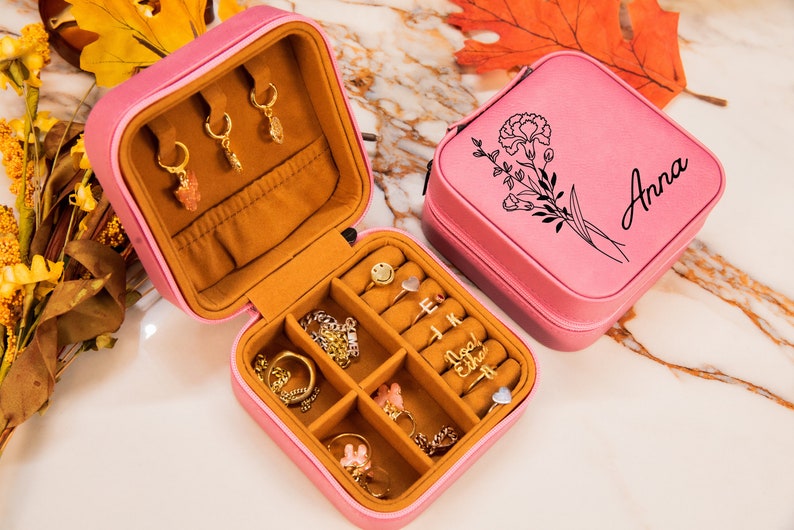 Puede incluir: Caja de joyer&iacute;a de terciopelo rosa con un dise&ntilde;o floral y el nombre "Anna" grabado en la tapa. La caja est&aacute; abierta y llena de varias piezas de joyer&iacute;a, incluyendo anillos, aretes y un collar.