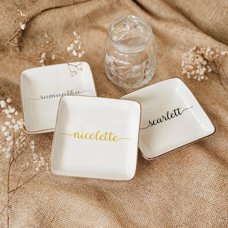 May include: Three white ceramic ring dishes with gold trim. The dishes are square and have the names "Samantha", "Nicolette", and "Scarlett" written in cursive on them.