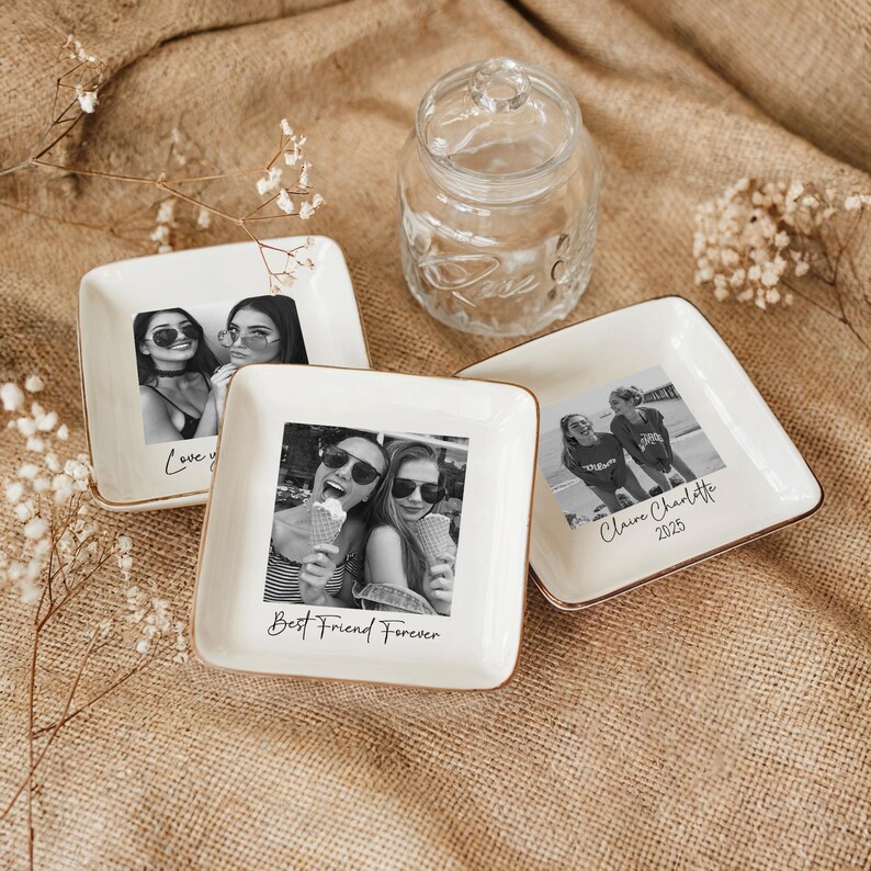 May include: White square ceramic trinket dishes with gold trim, each featuring a black and white photograph. One dish reads "Love you", another "Best Friend Forever", and a third "Claire Charlotte 2025". A clear glass jar is in the background.