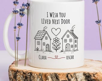 May include: White ceramic mug with the text "I Wish You Lived Next Door" and a house illustration. The mug features the names "Clara" and "Oscar" below the houses, connected by a heart. The mug is displayed on a wooden slice.