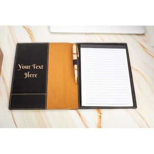 May include: A black leather portfolio with a brown leather flap and a pen holder. The portfolio has a lined notepad inside and the text "Your Text Here" is engraved on the front.