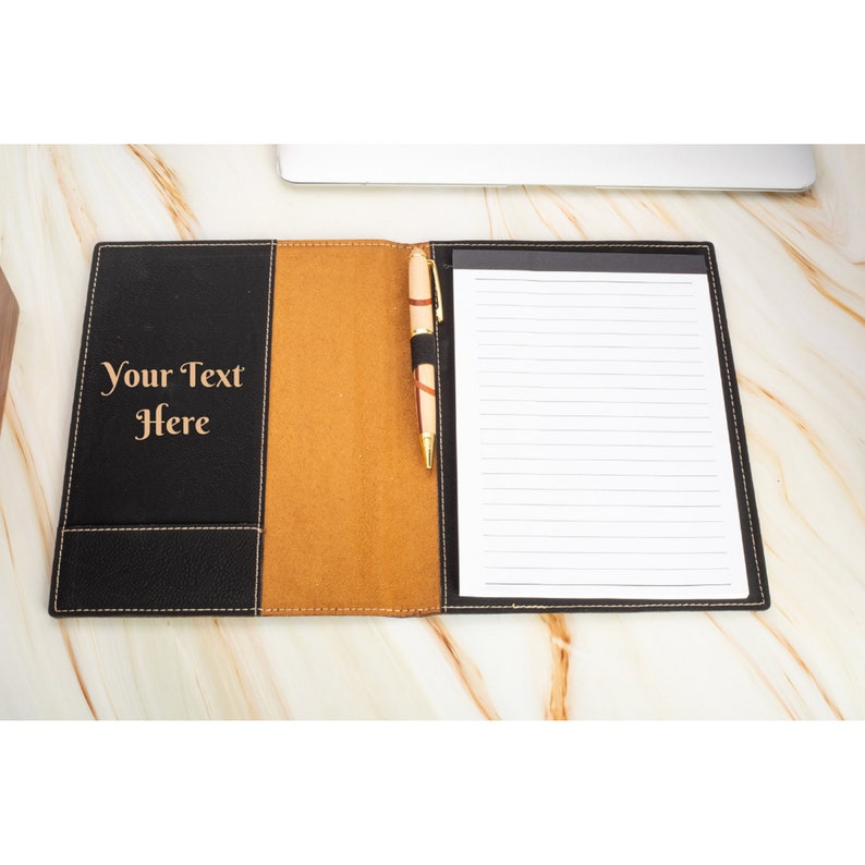 May include: A black leather portfolio with a brown leather flap and a pen holder. The portfolio has a lined notepad inside and the text "Your Text Here" is engraved on the front.