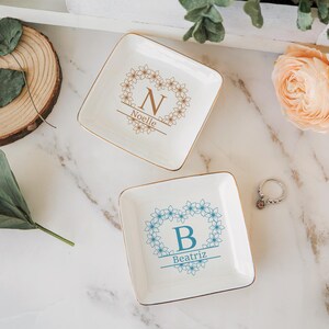 Op de afbeelding: Twee vierkante, witte keramische ringschalen met gouden rand. E&eacute;n schaal heeft een bloemenkrans met de letter "N" en de naam "Noelle". De andere heeft een blauwe bloemenkrans met de letter "B" en de naam "Beatriz". Er is ook een zilveren ring aanwezig.