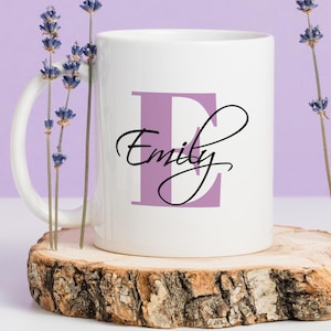 May include: White ceramic mug with a purple letter E and the name "Emily" written in cursive. The mug is sitting on a wooden coaster.