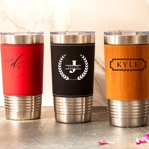 May include: Three stainless steel tumblers with personalized leather wraps. The tumblers are red, black, and brown. The red tumbler has the name "Daisy" on it. The black tumbler has a wreath with the letter "J" and the name "Jalen" on it. The brown tumbler has the name "Kyle" on it.