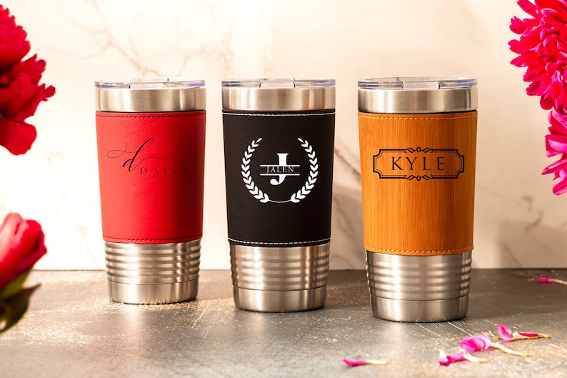 May include: Three stainless steel tumblers with personalized leather wraps. The tumblers are red, black, and brown. The red tumbler has the name "Daisy" on it. The black tumbler has a wreath with the letter "J" and the name "Jalen" on it. The brown tumbler has the name "Kyle" on it.