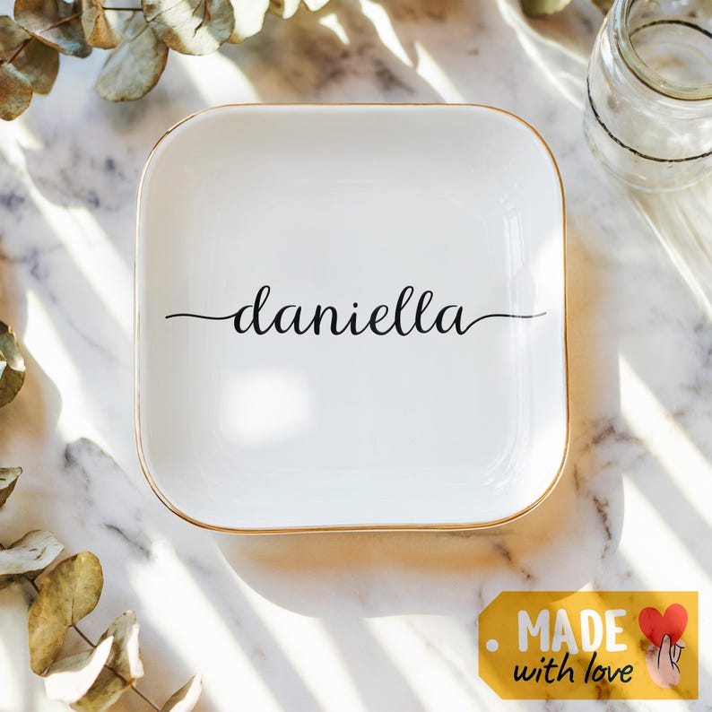 May include: White ceramic trinket tray with a gold rim. The tray has the name "daniella" written in cursive on it.