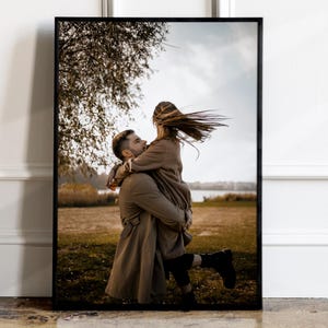May include: A couple in love, the man is holding the woman in his arms, they are both wearing brown coats, the woman's hair is blowing in the wind, they are standing in a field with trees and a lake in the background.