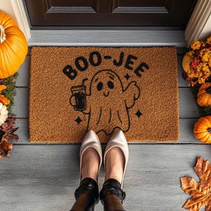 May include: A brown coir doormat with the text "BOO-JEE" in black lettering. A cartoon ghost holding a cup of coffee is in the center of the mat.