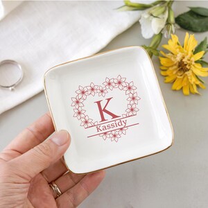 Op de afbeelding: Een vierkant, cr&egrave;mekleurig keramisch sieradenschaaltje met een gouden rand. Het schaaltje heeft een bloemenhartontwerp met de letter "K" en de naam "Kassidy" in rood. Een ring en bloemen zijn op de achtergrond.