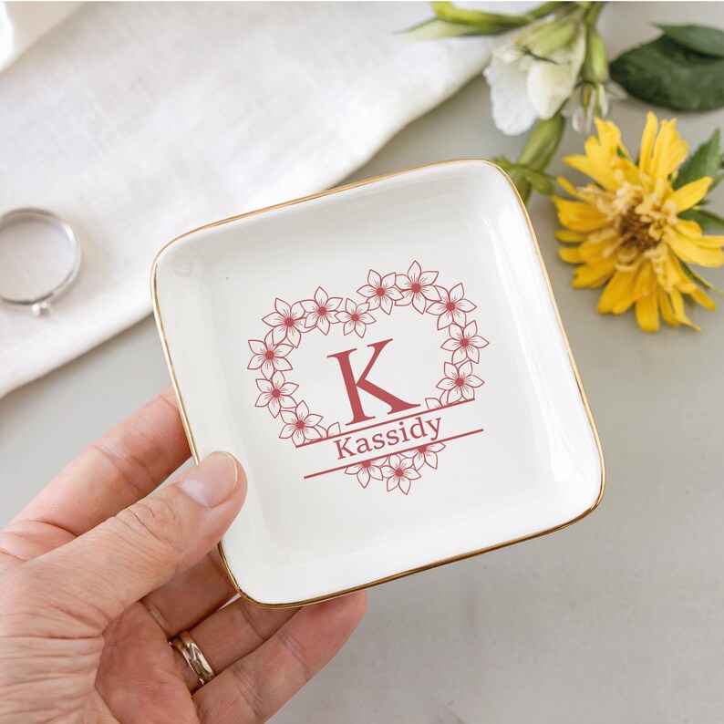 Op de afbeelding: Een vierkant, cr&egrave;mekleurig keramisch sieradenschaaltje met een gouden rand. Het schaaltje heeft een bloemenhartontwerp met de letter "K" en de naam "Kassidy" in rood. Een ring en bloemen zijn op de achtergrond.