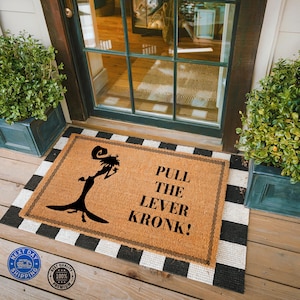 May include: A rectangular doormat with the text "PULL THE LEVER KRONK!" in black. The mat has a brown center with a black and white striped border. A black silhouette of a character is on the left side.