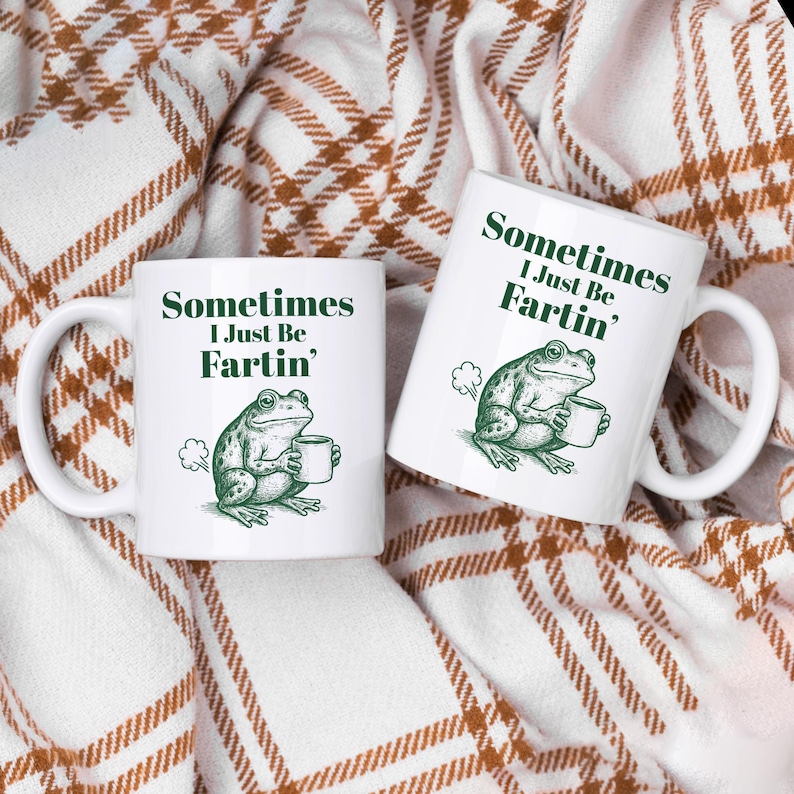 May include: Two white ceramic mugs with the text "Sometimes I Just Be Fartin'" and a green illustration of a frog holding a mug. The mugs are on a brown and white plaid blanket.