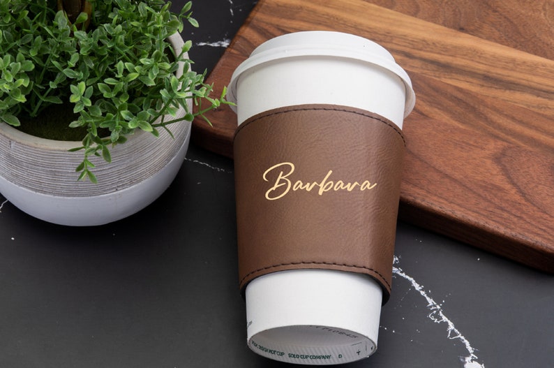Puede incluir: Una funda de taza de caf&eacute; de cuero marr&oacute;n con el nombre "Barbara" escrito en letras doradas. La funda est&aacute; en una taza de caf&eacute; para llevar blanca.