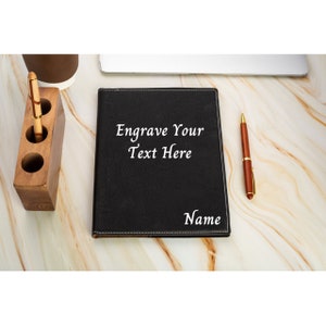 May include: A black leather journal with white stitching and the text "Engrave Your Text Here" and "Name" on the front cover.