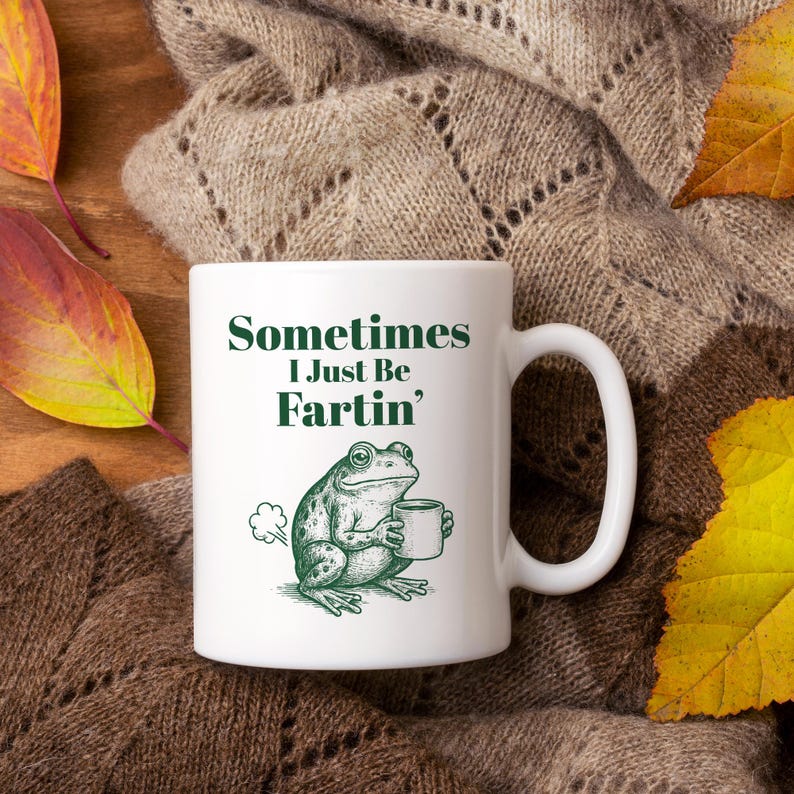 May include: White ceramic mug with the text "Sometimes I Just Be Fartin'" in green. A green frog illustration holding a mug is on the front. The mug is set on a brown knitted blanket with autumn leaves.