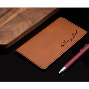 May include: A brown leather checkbook cover with the name "Anthony Hall" engraved in black lettering. The cover is lying on a black surface next to a burgundy pen.
