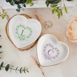 Op de afbeelding: Twee hartvormige ringhouders met gouden rand. E&eacute;n schaal heeft een groen bloemmotief met de letter "L" en de naam "Let&iacute;cia". De andere schaal heeft een paars bloemmotief met de letter "C" en de naam "Carolina".