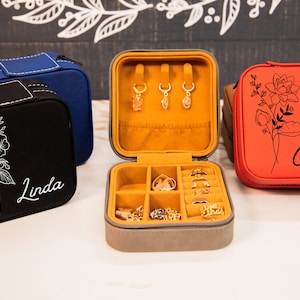 Puede incluir: Tres cajas de joyer&iacute;a cuadradas con exteriores de diferentes colores y nombres personalizados. Las cajas est&aacute;n cerradas, excepto la caja del medio, que est&aacute; abierta para revelar un interior de terciopelo amarillo con compartimentos para anillos y pendientes.