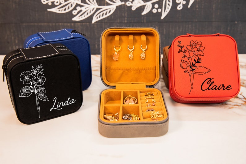Puede incluir: Tres cajas de joyer&iacute;a cuadradas con exteriores de diferentes colores y nombres personalizados. Las cajas est&aacute;n cerradas, excepto la caja del medio, que est&aacute; abierta para revelar un interior de terciopelo amarillo con compartimentos para anillos y pendientes.