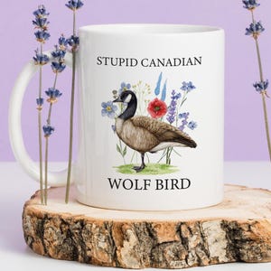 May include: White ceramic mug featuring a watercolor illustration of a Canada goose with floral accents. The mug has the text "STUPID CANADIAN" and "WOLF BIRD" printed on it. The mug is displayed on a wooden slice.