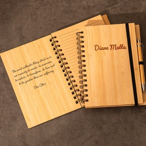 May include: A bamboo notebook with a spiral binding, open to reveal lined pages and a quote. The cover is engraved with the name "Diane Malla". A matching pen is included, held by a black elastic band. The notebook is on a dark gray surface.