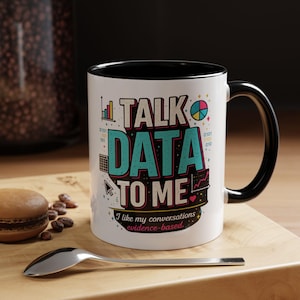 May include: A white ceramic mug with a black interior and handle, featuring the text "TALK DATA TO ME" in large, colorful letters, along with "I like my conversations evidence-based." and various small data-themed icons like charts and binary code.