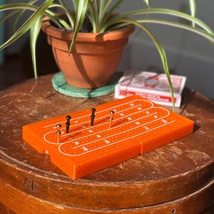 Puede incluir: Un tablero de cribbage naranja con clavijas negras y plateadas se muestra sobre una mesa de madera. El tablero tiene líneas blancas y números que marcan los caminos de puntuación. Un mazo de cartas y una planta en maceta están en el fondo.