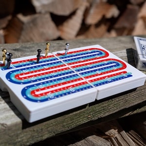 May include: A white plastic cribbage board featuring two parallel tracks with red and blue printed numbering from start to 120. Four metal pegs in gold, silver, and black are inserted into the board. A deck of playing cards is visible nearby.