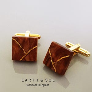 May include: A pair of square cufflinks with a wood grain appearance. The cufflinks are brown with gold accents and gold-colored clasps. The text "EARTH & SOL Handmade in England" is at the bottom.
