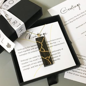 May include: A rectangular dark wood pendant necklace featuring gold-coloured Kintsugi-style cracks, presented in a black gift box with a white ribbon. The set includes informational cards detailing the Kintsugi philosophy and the use of ancient bog oak.