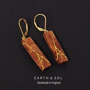 May include: A pair of rectangular wooden earrings with gold-colored accents. The earrings have a gold-colored hook closure. The earrings are brown with a gold-colored design. The text "EARTH & SOL Handmade In England" is at the bottom.
