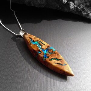 May include: A wooden pendant necklace with a silver chain. The pendant is a light brown, elongated oval shape with blue and green accents. The pendant has a natural wood grain pattern and a polished finish.