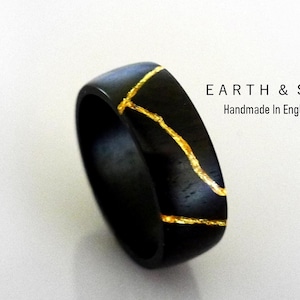 May include: A black ring featuring a jagged, metallic gold-colored vein running across its surface. The text EARTH & SOL Handmade In England is printed in black to the right of the ring against a plain, light-colored background.