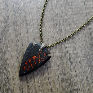 Op de afbeelding: Een ketting met een zwarte pijlpunt hanger met een rood en oranje ontwerp. De ketting is gemaakt van hout en heeft een bronzen ketting.
