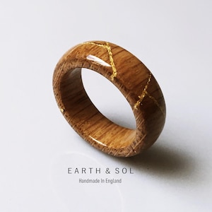 May include: A wooden ring with a natural wood grain finish and gold-colored inlay. The ring is a band style and features the text "EARTH & SOL Handmade In England".