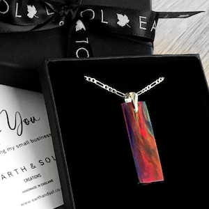 Könnte beinhalten: Eine silberne Kettenkette mit einem rechteckigen Anhänger aus buntem Harz. Der Anhänger hat ein wirbelndes Muster aus Rot, Orange, Gelb, Grün, Blau und Lila. Die Kette befindet sich in einer schwarzen Geschenkbox mit einem schwarzen Band und einem Etikett mit der Aufschrift "Danke" und "Earth & Sol Creations".