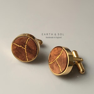 Puede incluir: Un par de gemelos redondos con un marco de metal dorado pulido y una incrustación de madera marrón circular con detalles de vetas doradas. El texto EARTH & SOL Handmade In England está impreso en negro sobre los gemelos.
