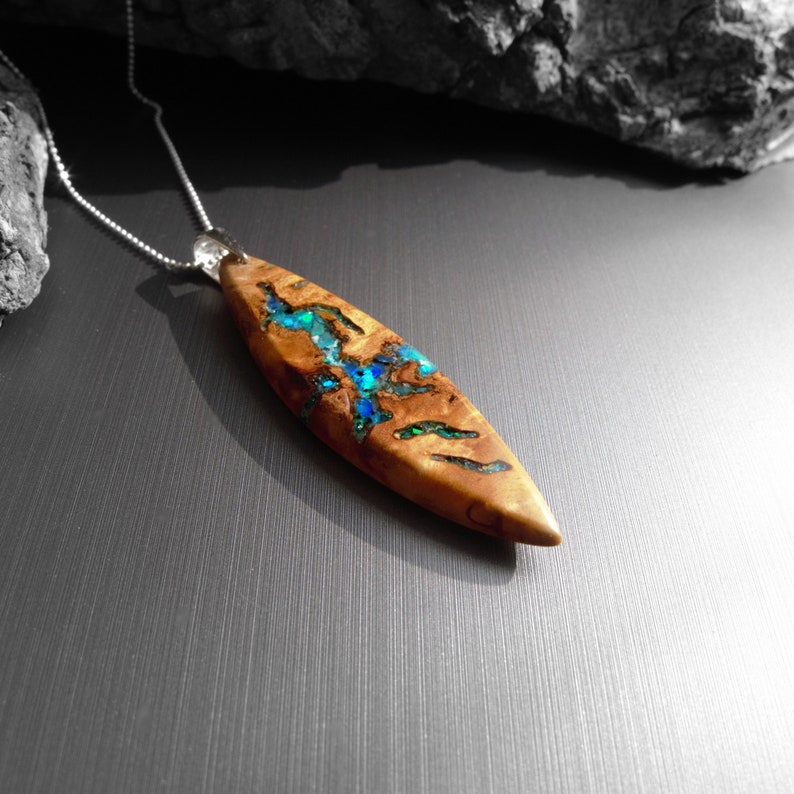May include: A wooden pendant necklace with a silver chain. The pendant is a light brown, elongated oval shape with blue and green accents. The pendant has a natural wood grain pattern and a polished finish.