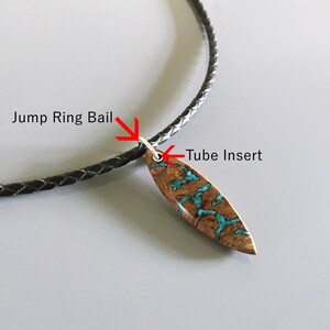 Puede incluir: Un collar con un colgante de madera marr&oacute;n en forma de tabla de surf con incrustaciones de color turquesa. El colgante est&aacute; unido a un cord&oacute;n trenzado negro mediante una anilla plateada y un inserto de tubo, con las etiquetas Jump Ring Bail y Tube Insert.