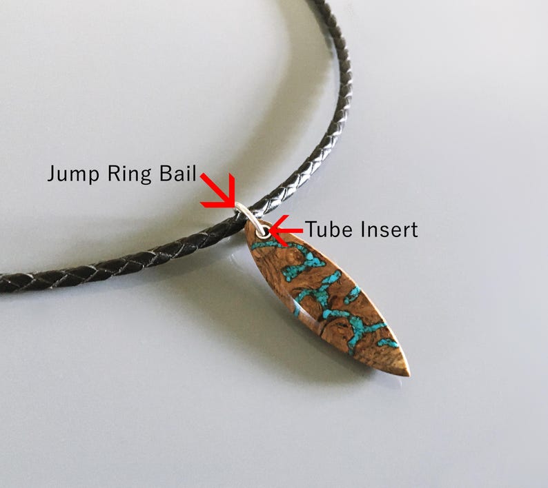 Puede incluir: Un collar con un colgante de madera marr&oacute;n en forma de tabla de surf con incrustaciones de color turquesa. El colgante est&aacute; unido a un cord&oacute;n trenzado negro mediante una anilla plateada y un inserto de tubo, con las etiquetas Jump Ring Bail y Tube Insert.