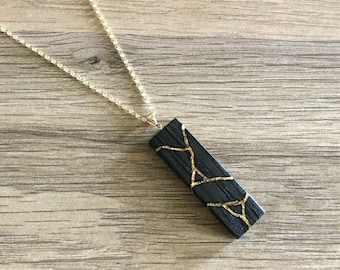 Puede incluir: Un colgante de madera negro con detalles dorados que cuelga de una cadena dorada. El colgante es rectangular y tiene un diseño único de líneas doradas.