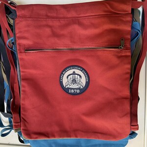 Op de afbeelding: Rode canvas trekkoordtas met een ritsvak en een ronde patch met een vuurtoren en de tekst "Cape Blanco Light Station 1870".