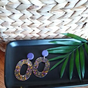 May include: A pair of gold and purple resin earrings with a geometric oval shape. The earrings have a purple clay bead at the top.
