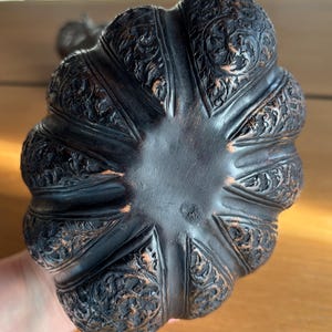 May include: A dark brown, intricately carved decorative object with a scalloped edge. The object features detailed floral and scrollwork patterns. The base is a smooth, dark brown color. The object appears to be made of wood.