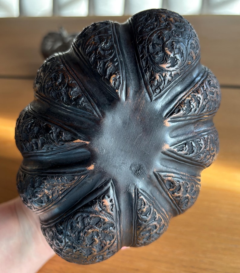 May include: A dark brown, intricately carved decorative object with a scalloped edge. The object features detailed floral and scrollwork patterns. The base is a smooth, dark brown color. The object appears to be made of wood.