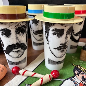 May include: A set of vintage drinking glasses with a black and white portrait of a man with a mustache. Each glass is topped with a small hat in various colours, including yellow, green, blue, and red. Two wooden toys with red balls are in the foreground.
