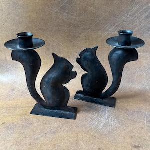 May include: Two black metal candle holders. Each holder features a stylized cat silhouette with a curved tail and a circular platform to hold a candle. The holders are on a rectangular base.